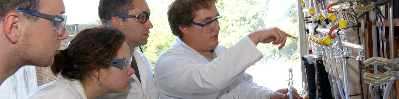 4 students in white lab coats and blue goggles watching a demonstration