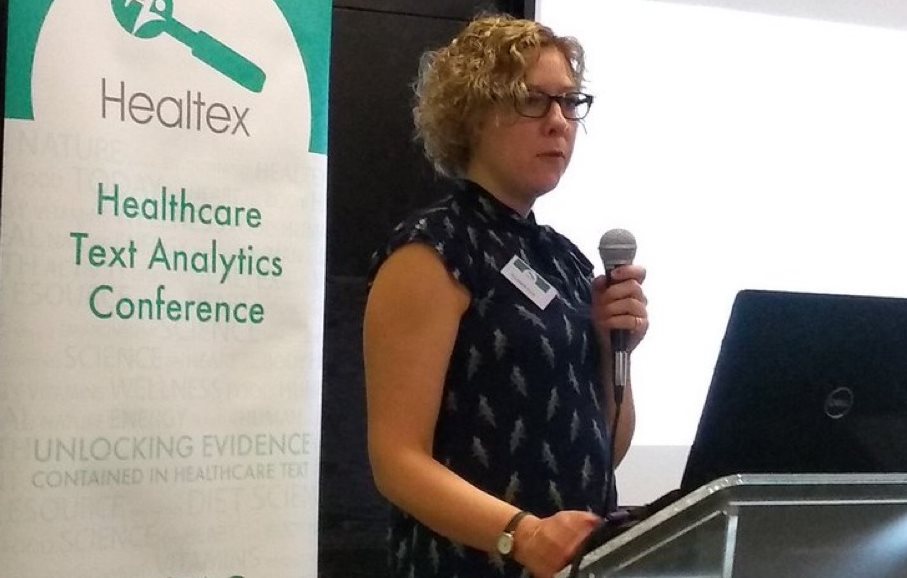 Dr Liz Ford standing with a laptop and microphone in front of some slides at the Healtex Healthcare Text Analytics Conference