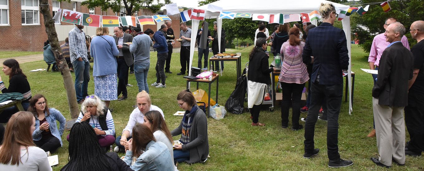 A photograph from an RNA event, showing bunting with flags of the world, with people sitting on the grass eating.