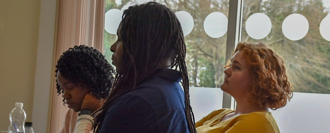 three people sat around a table listening to a speaker. one is making notes