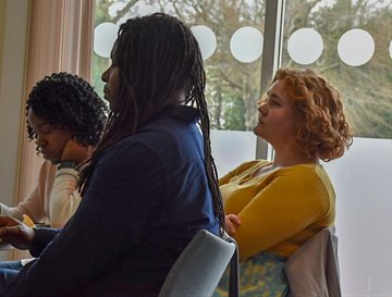 three people sat around a table listening to a speaker. one is making notes