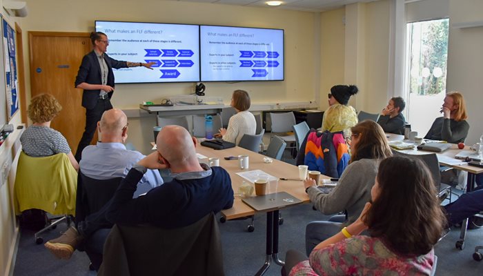 A room full of people listening to a man presenting. the man is pointing to a tv screen