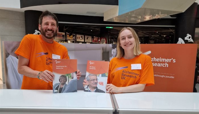 Two researchers Ben Hicks and Kat Wheatley wearing orange t shirts stood on a stall in a shopping centre promoting Alzheimers Research