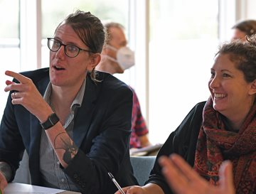 A man with glasses pointing and a woman sat next to him smiling. Both are looking ahead