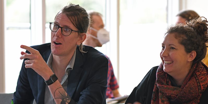 A man with glasses pointing and a woman sat next to him smiling. Both are looking ahead