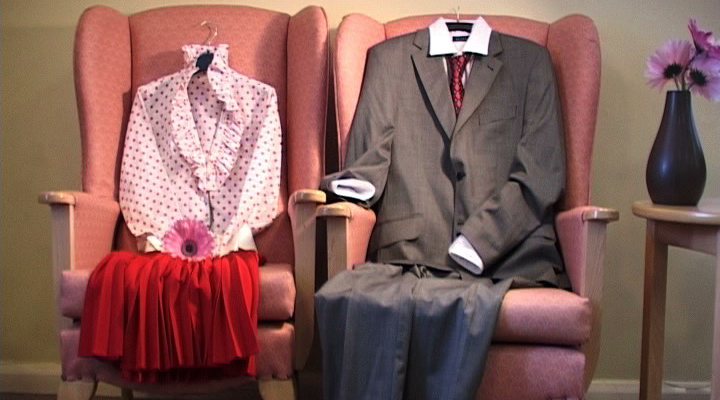 two faded pink arm chairs, with 2 sets of empty clothes arranged as if a person was sitting in them.