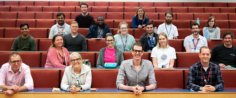 A photo showing a group of researchers sat on red seats in a lecture theatre smiling at the camera