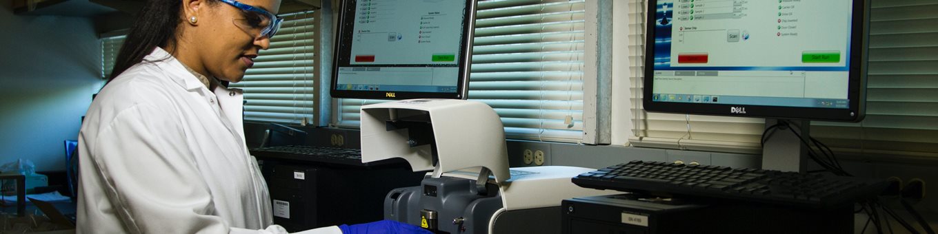 An image of a girl in a white lab coat and blue gloves carrying out an experiment with two computer screens in a medical laboratory