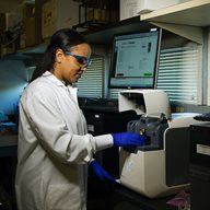 An image of a girl in a white lab coat and blue gloves carrying out an experiment with two computer screens in a medical laboratory