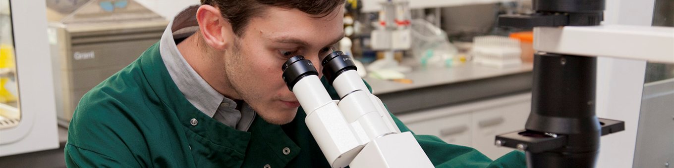 Research student in a green lab coat looking down a microscope