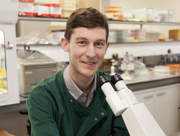 A man smiling wearing a green lab coat in front of a microscope