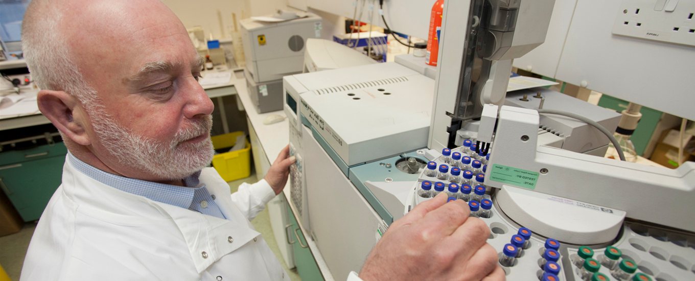 A researcher with a white beard and white lab coat selects a test tube