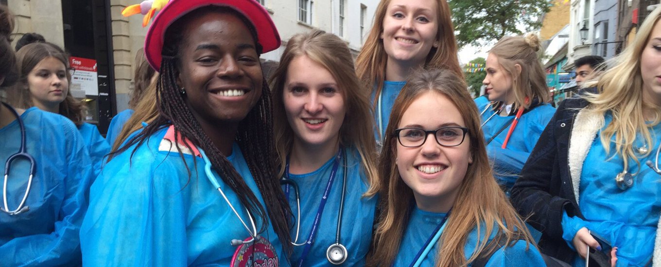 Smiling group of MedSoc students, in blue gowns and stethoscopes, on fundraising event