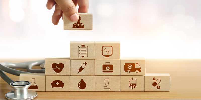 A hand putting wooden blocks on top of each other. Each block has a red illustration of a medical theme like a heart, ambulance and thermometer