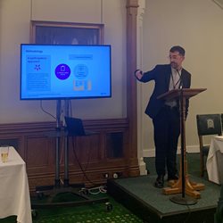 A lecturer named Diego Garcia, stood on a podium in front of a screen and green carpet at the House of Commons