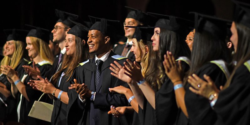 Lots of students stood wearing graduation gowns and hats facing forwards clapping