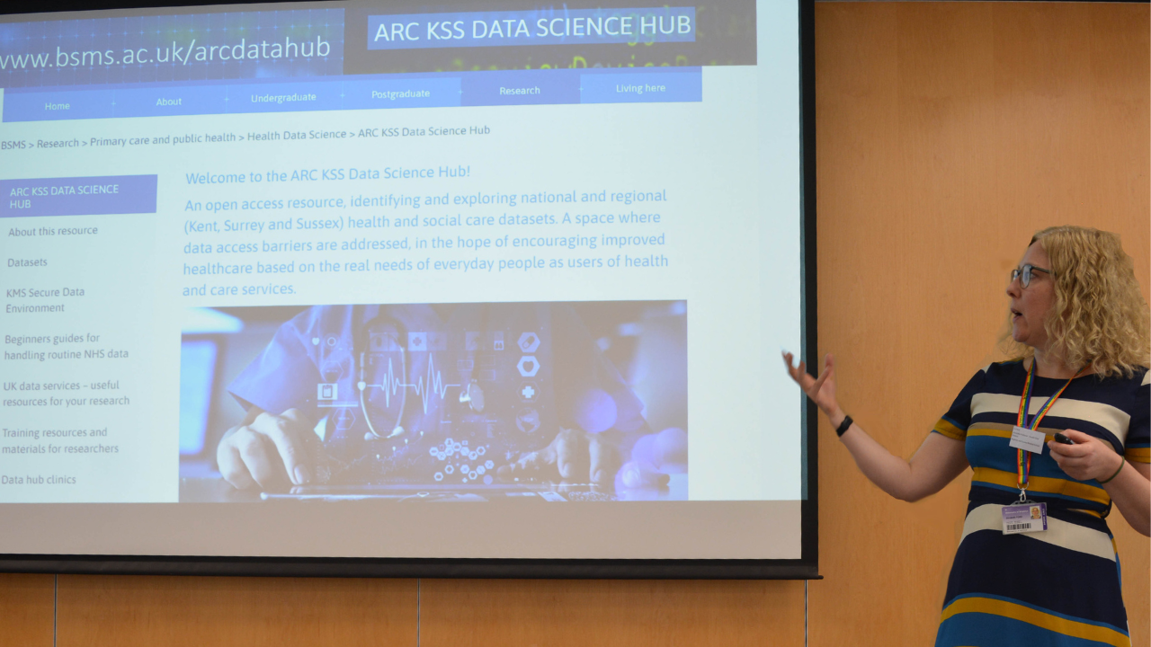Liz Ford standing next to a presentation screen in the lecture theatre with the ARC KSS Data Science Hub website up on the screen