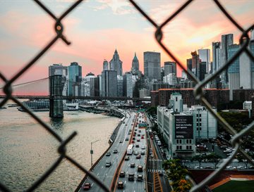 A photo showing a scenic skyline of buildings through a barbed wire fence with the sun setting. Cars and a road are also in the shot