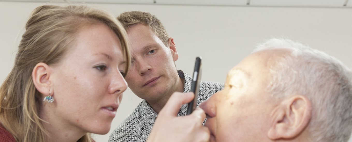 Female student examines eyes of older male patient educator, watched by male teacher