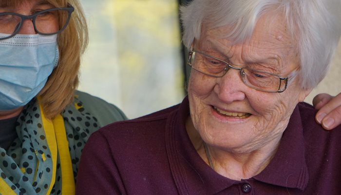 A carer with a mask on and an elderly woman smiling with an arm around her shoulder