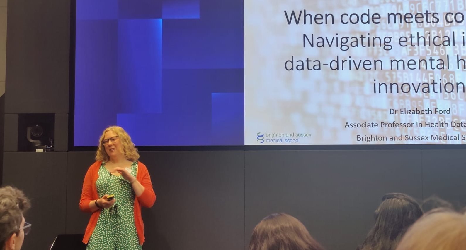Liz standing at the front of an audience with a microphone in front of a screen giving a presentation