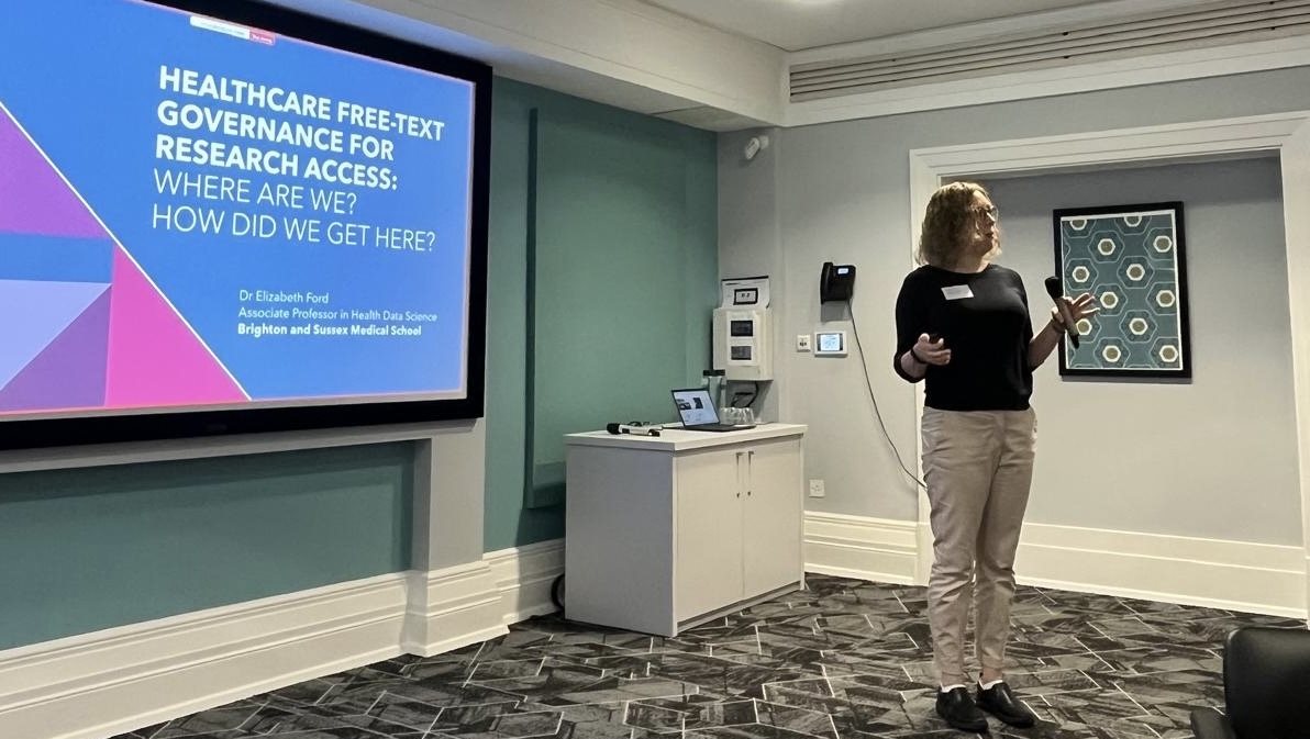 Liz Ford standing at the front of a room in front of a large screen presenting a presentation at Safetext event