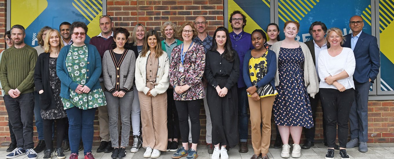 A group photo of the primary care team at BSMS. The team are stood in front of a wall with bright artwork on it