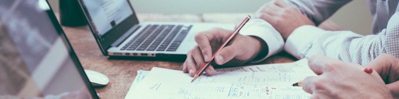 Two people working from two laptops, with just their arms and hands in view. One person is also making notes on a notepad