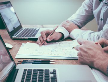 Two people working from two laptops, with just their arms and hands in view. One person is also making notes on a notepad