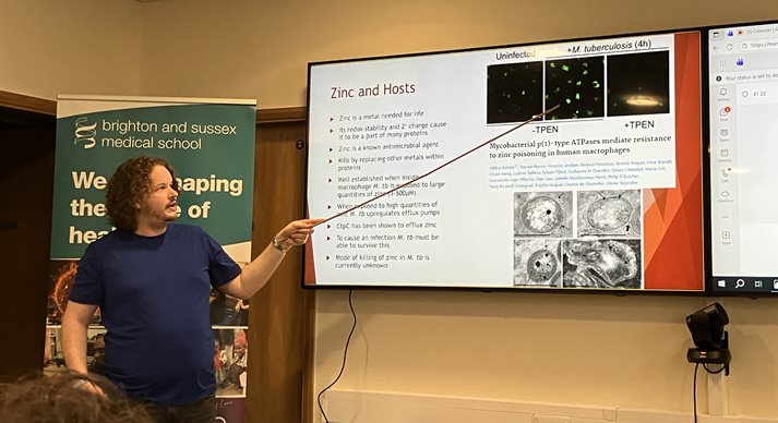 Someone at the front of a seminar room pointing at the board whilst giving a presentation