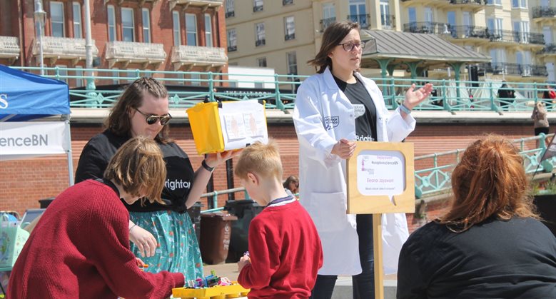 Someone stood in a lab coat giving a demonstrating whilst others watch on the beach