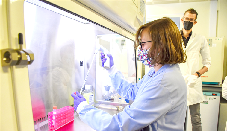Two people stood in a lab, one of them holding a test tube and beaker and the other observing