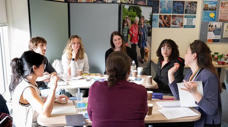 A group of people sat around a table in a seminar room having a discussion with papers and books in front of them