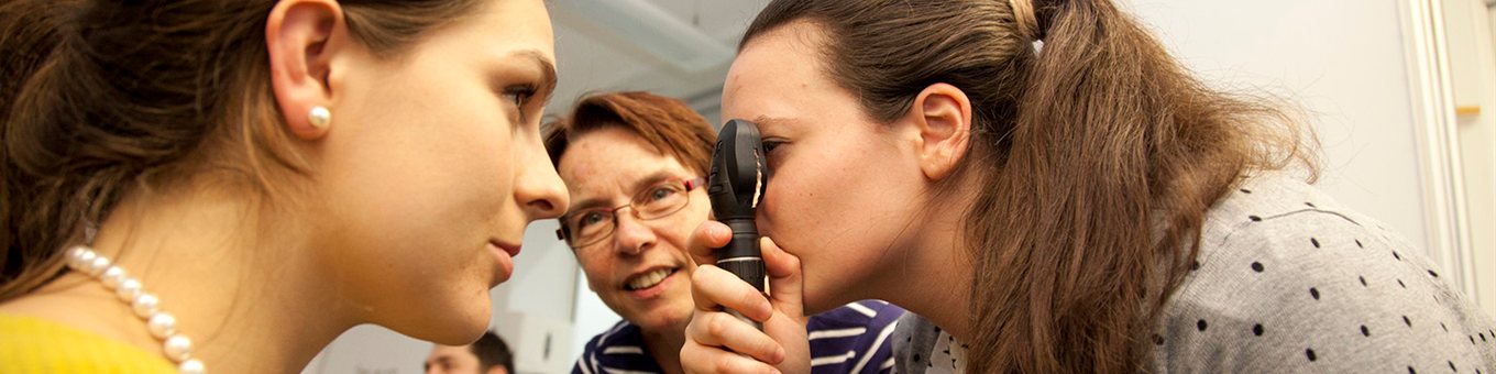 A student examines another's eye, watched by a teacher