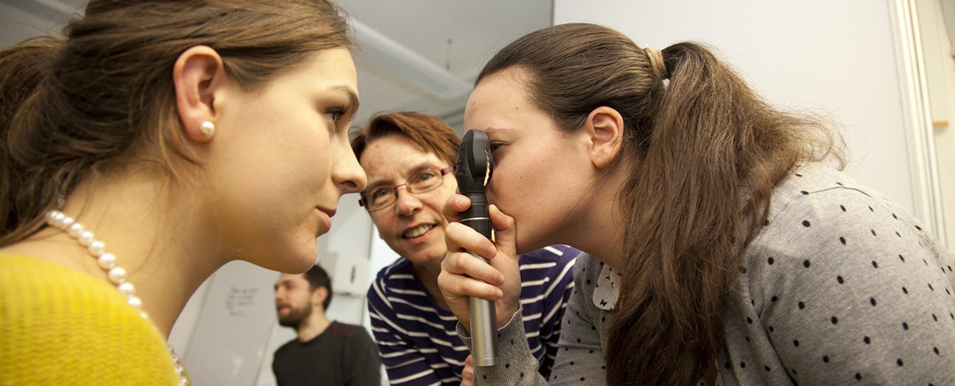 A female student uses an ophthalmic torch to examine another's eyes, looked on by teacher