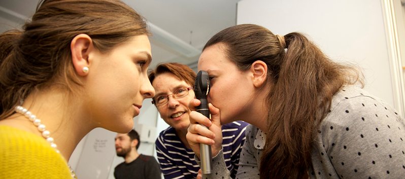 A female student examines another's eye, using a torch, while the teacher looks on