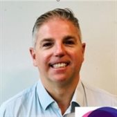 Portrait photograph of Damian Brewer, a BSMS Public Health alumnus, who is wearing a light blue shirt and smiling at the camera
