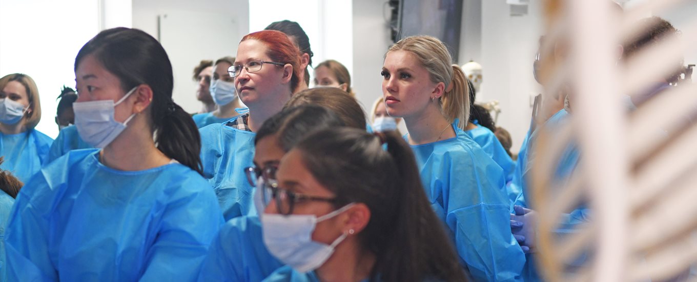 Students in blue scrubs and face masks listening to a talk in an anatomy lab