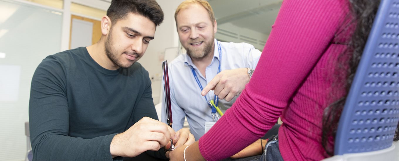 A male student tests a female students reflexes, watched by teacher