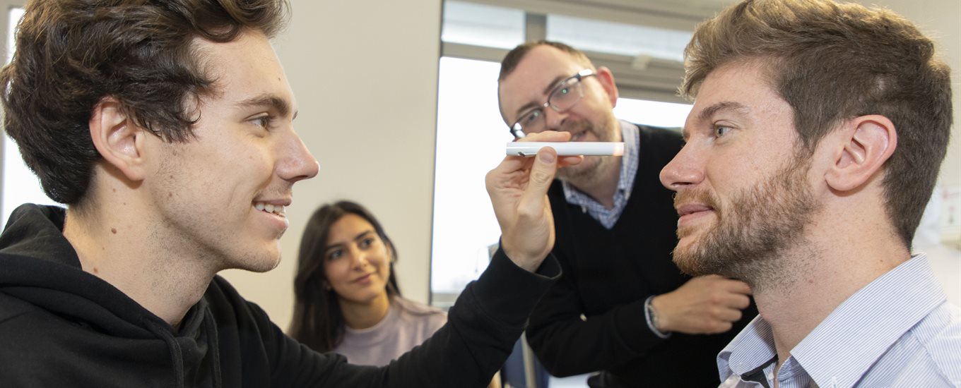 A male student shines a light into another student's eyes, watched on by teacher