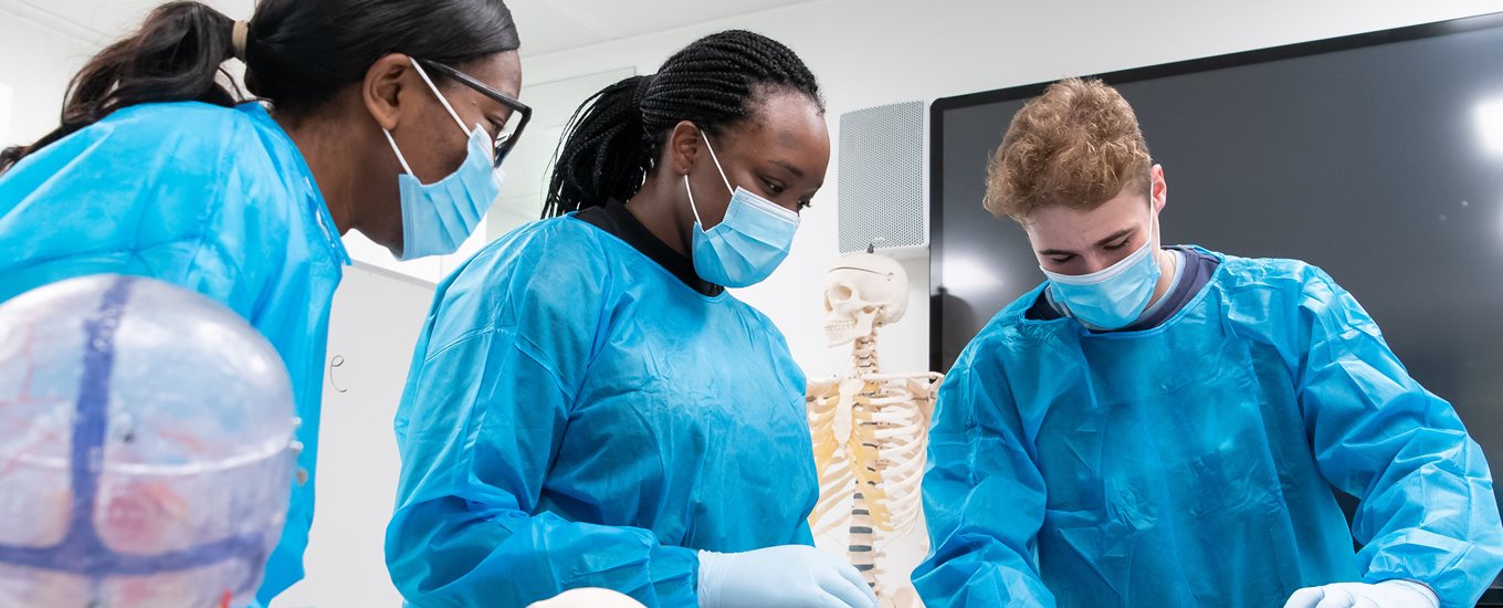 three bsms students in blue scrubs with plastic anatomical models in front of a screen