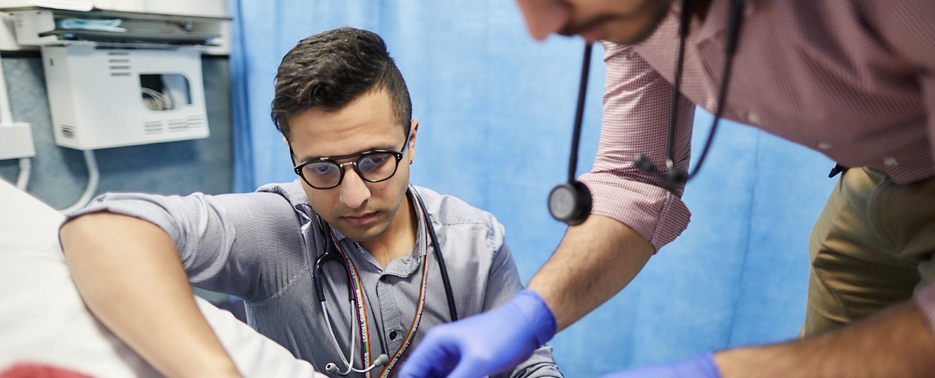 A student inserts a cannula into a patient's arm, watched on by another student