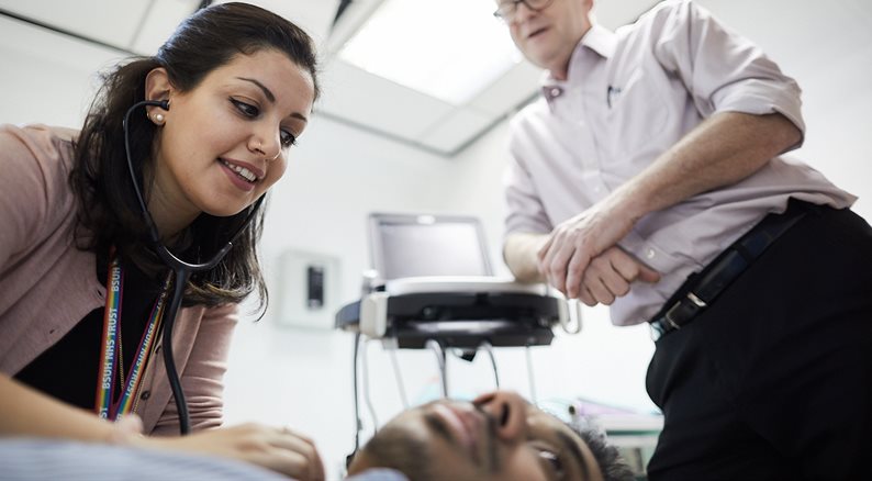 Female student using stethascope on a patient, overseen by a consultant