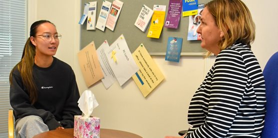 A female student sat on a chair talking to a woman in a blue chair with a table in between them