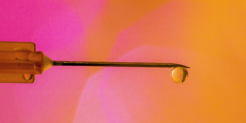 A close up of a needle with a drop of fluid on a colourful pink and orange background