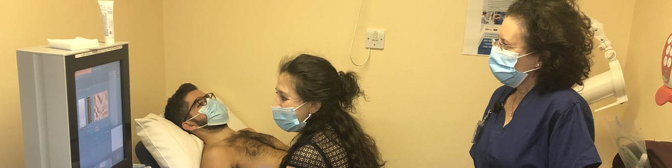 A clinician named Sumita Verma carrying out a fibroscan on a patient with a nurse, looking at a computer screen in a yellow room