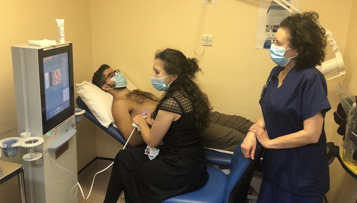 A clinician named Sumita Verma carrying out a fibroscan on a patient with a nurse, looking at a computer screen in a yellow room