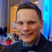 A head and shoulders shot of Andrew Barritt smiling at the camera wearing smart clothes inside a blue lit building