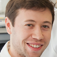 A close up shot of a man with brown hair, smiling, wearing a white lab coat