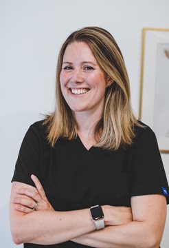 A woman named Charlotte Thompson standing with her arms folded, smiling at the camera in front of a photo frame and wearing a black top and smart watch